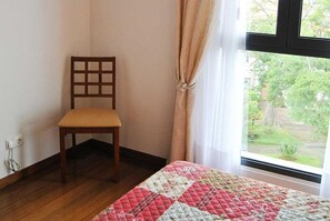 Una habitación con piso de madera, una silla, una cama con colcha y una ventana con cortinas, mostrando vistas de vegetación al exterior.