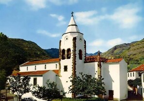 Una iglesia blanca con un campanario y un reloj, rodeada de vegetación y colinas.