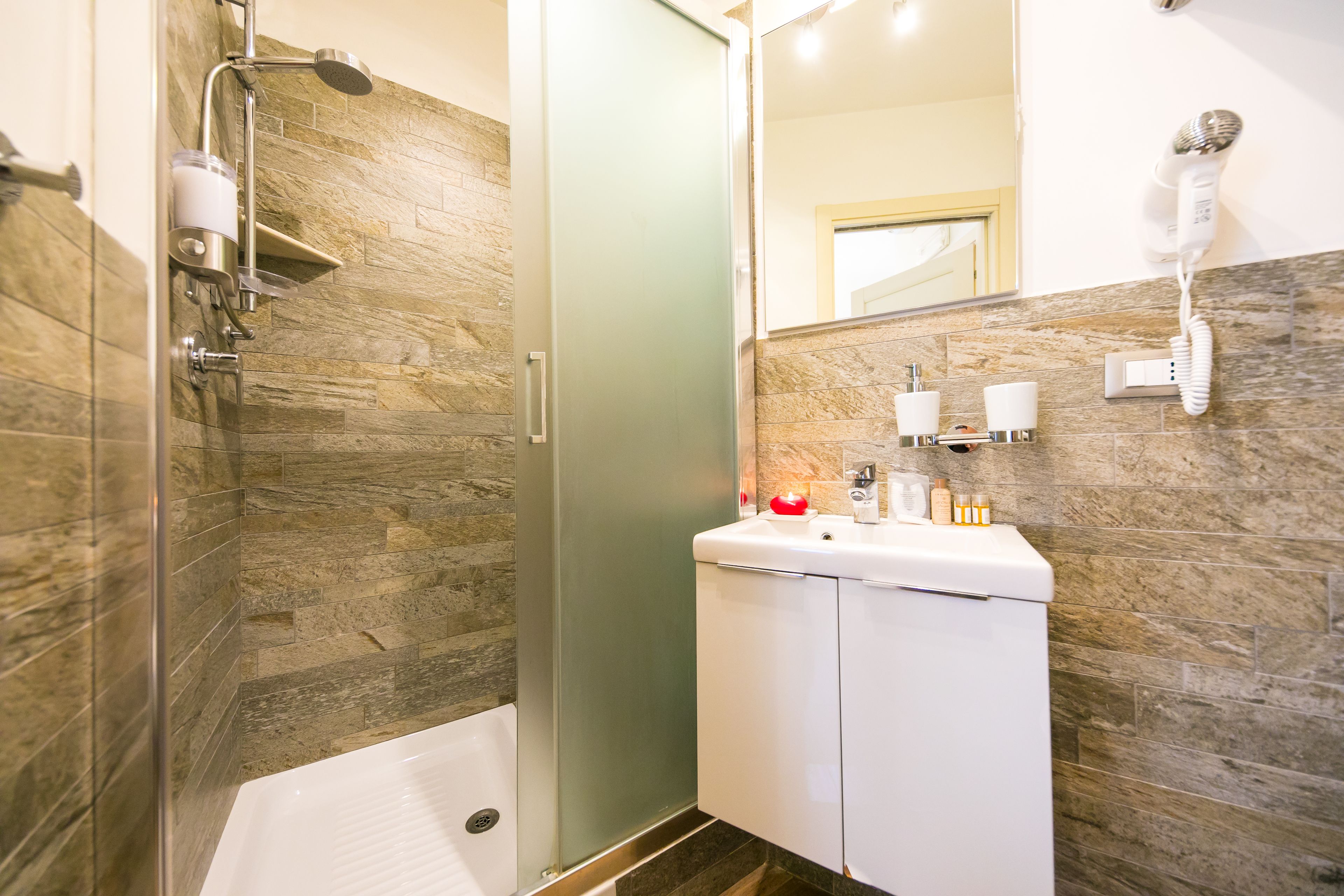 A modern bathroom with a glass shower enclosure, a white sink with toiletries, and a hairdryer mounted on the wall.