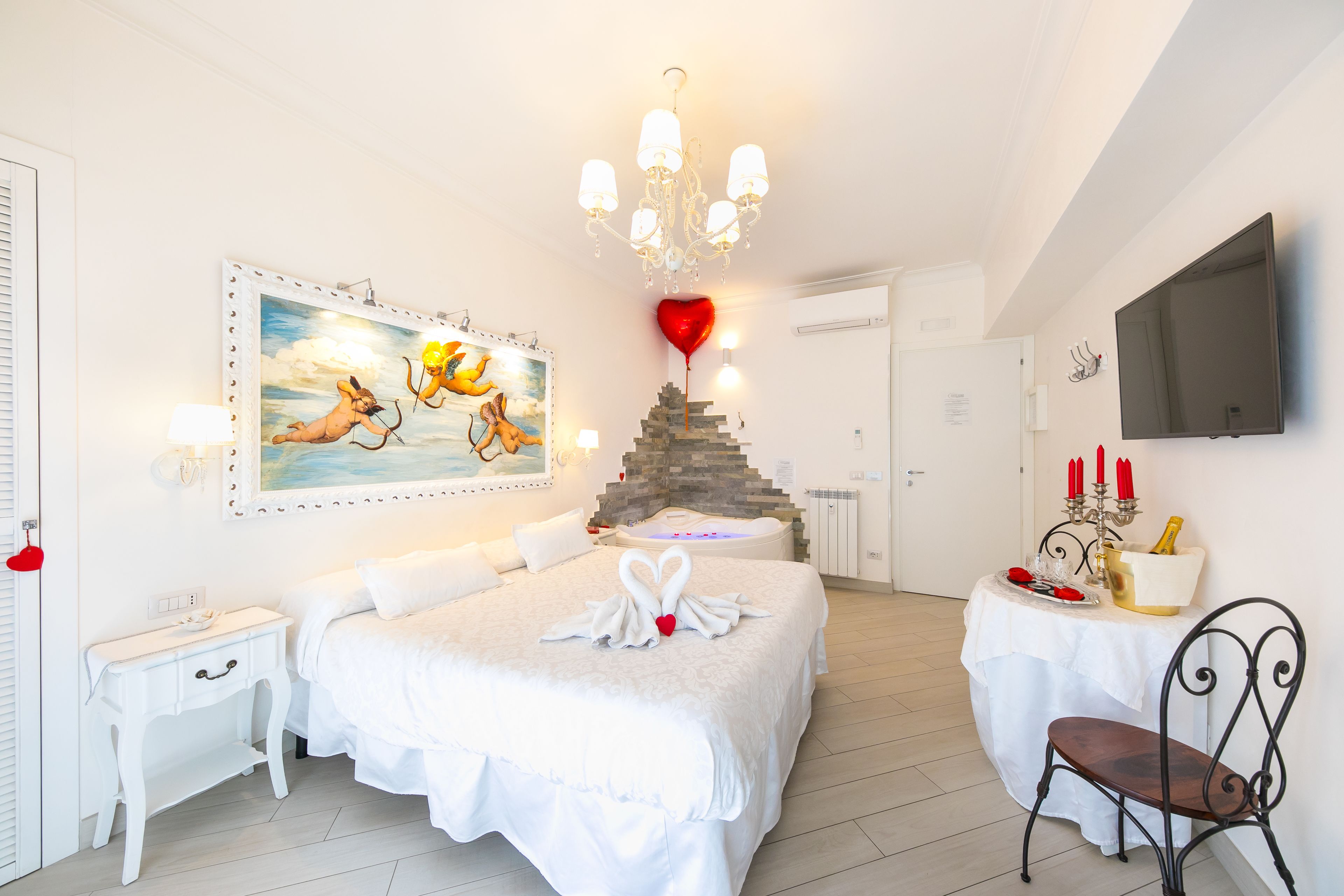 A bedroom with a large bed, a small table with a bottle and glasses, a chair, a painting on the wall, and a chandelier.