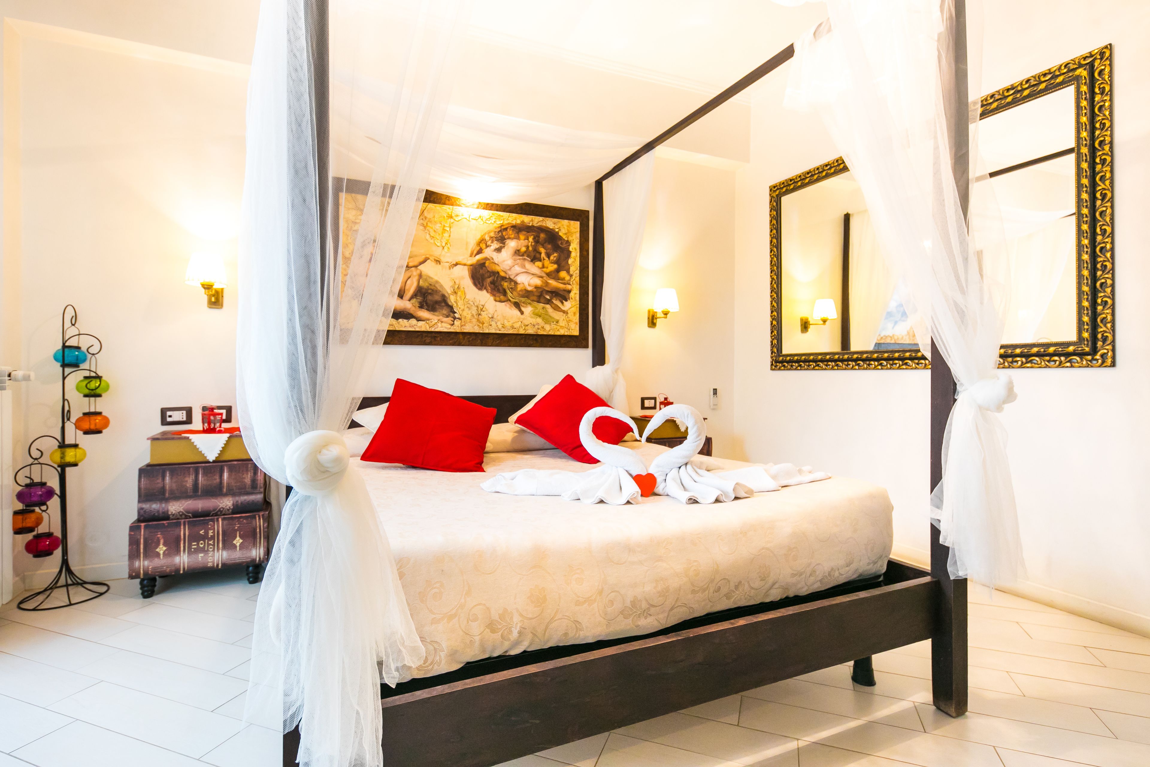 A four-poster bed with a canopy, red pillows, a painting on the wall, a mirror, and a bedside table with a lamp.