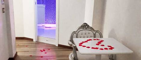 A hallway with a table set for two, adorned with rose petals, leading to a bathroom with a blue-lit shower area.