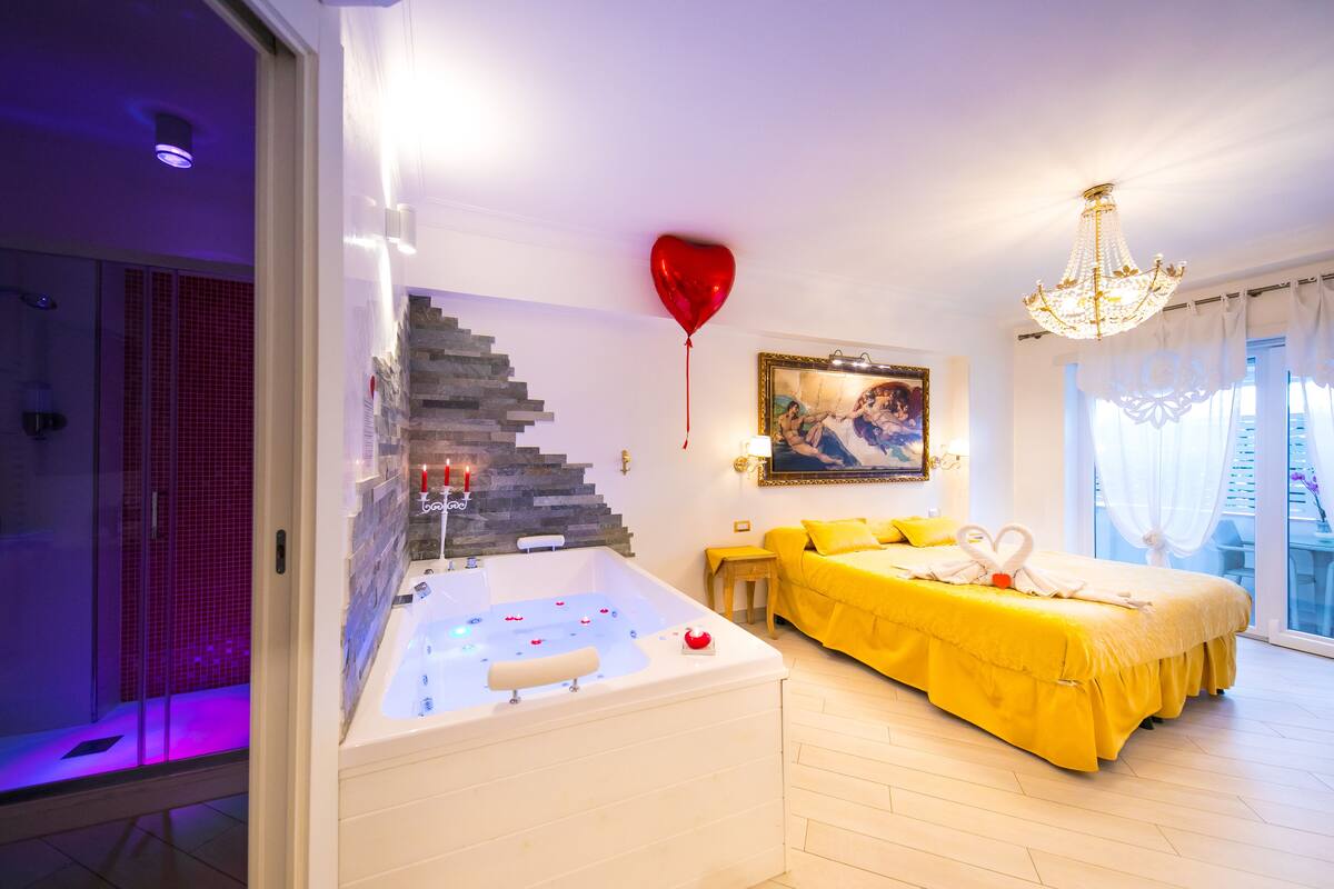 Hotellihuone, jossa on suuri kylpyamme, keltainen vuodevaatteilla varustettu sänky, seinällä taulu ja punainen sydämenmuotoinen ilmapallo.