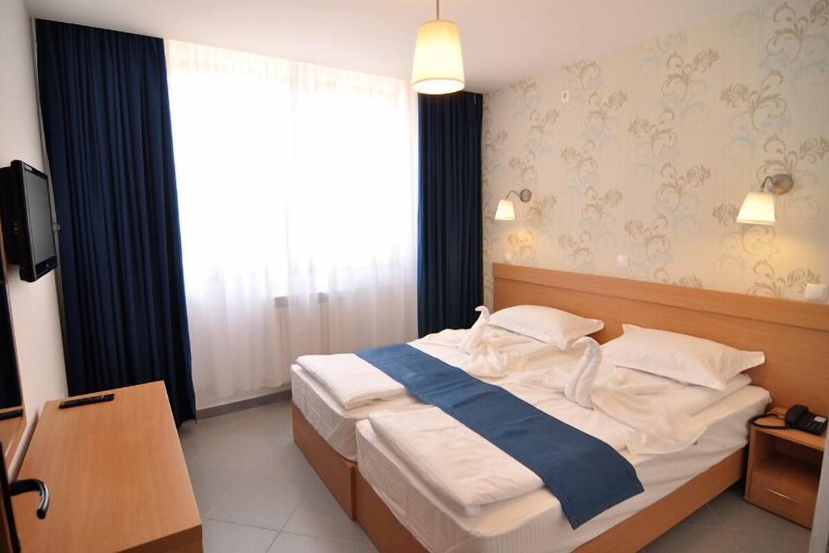 Ein Hotelzimmer mit Doppelbett, Schreibtisch, Stuhl, Fernseher und einem Fenster mit Vorhängen.