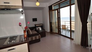 Un salon moderne avec un canapé, une table basse et un téléviseur. Il y a également un coin cuisine avec une cuisinière et une table à manger. La pièce est dotée de grandes portes vitrées qui donnent sur un balcon offrant une vue sur la mer.