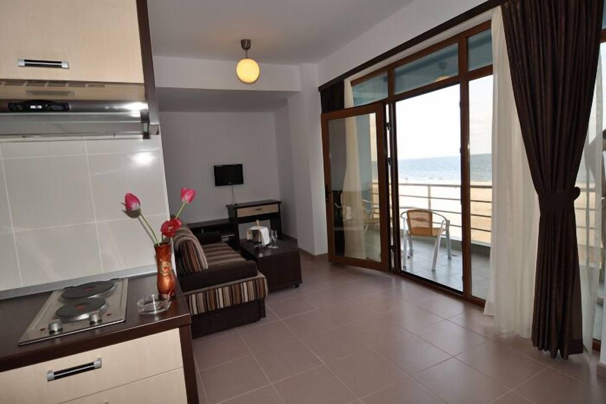 Ein modernes Wohnzimmer mit Sofa, Couchtisch und Fernseher. Es gibt eine Küchenzeile mit Herd und Esstisch. Der Raum verfügt über große Glasüren, die auf einen Balkon mit Meerblick führen.