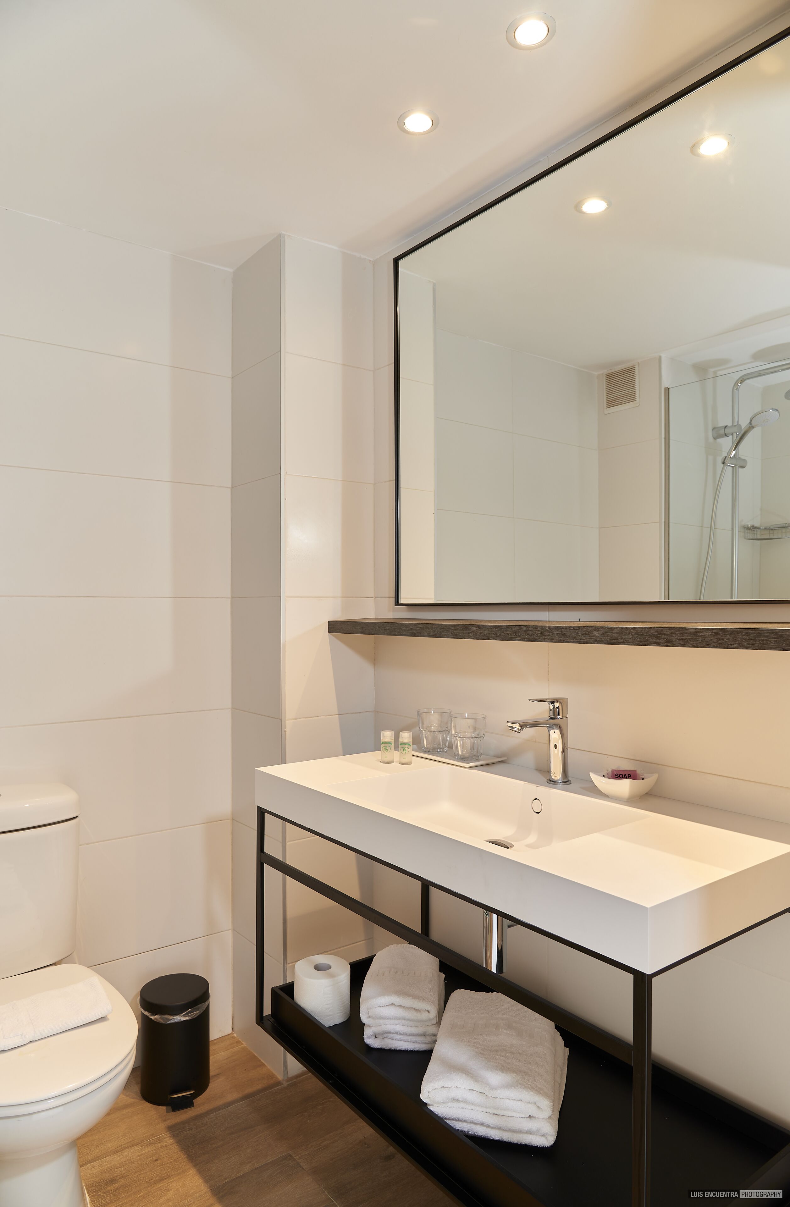 Un baño moderno con un lavamanos, espejo y retrete blancos. Hay toallas en un estante y un cesto de basura.