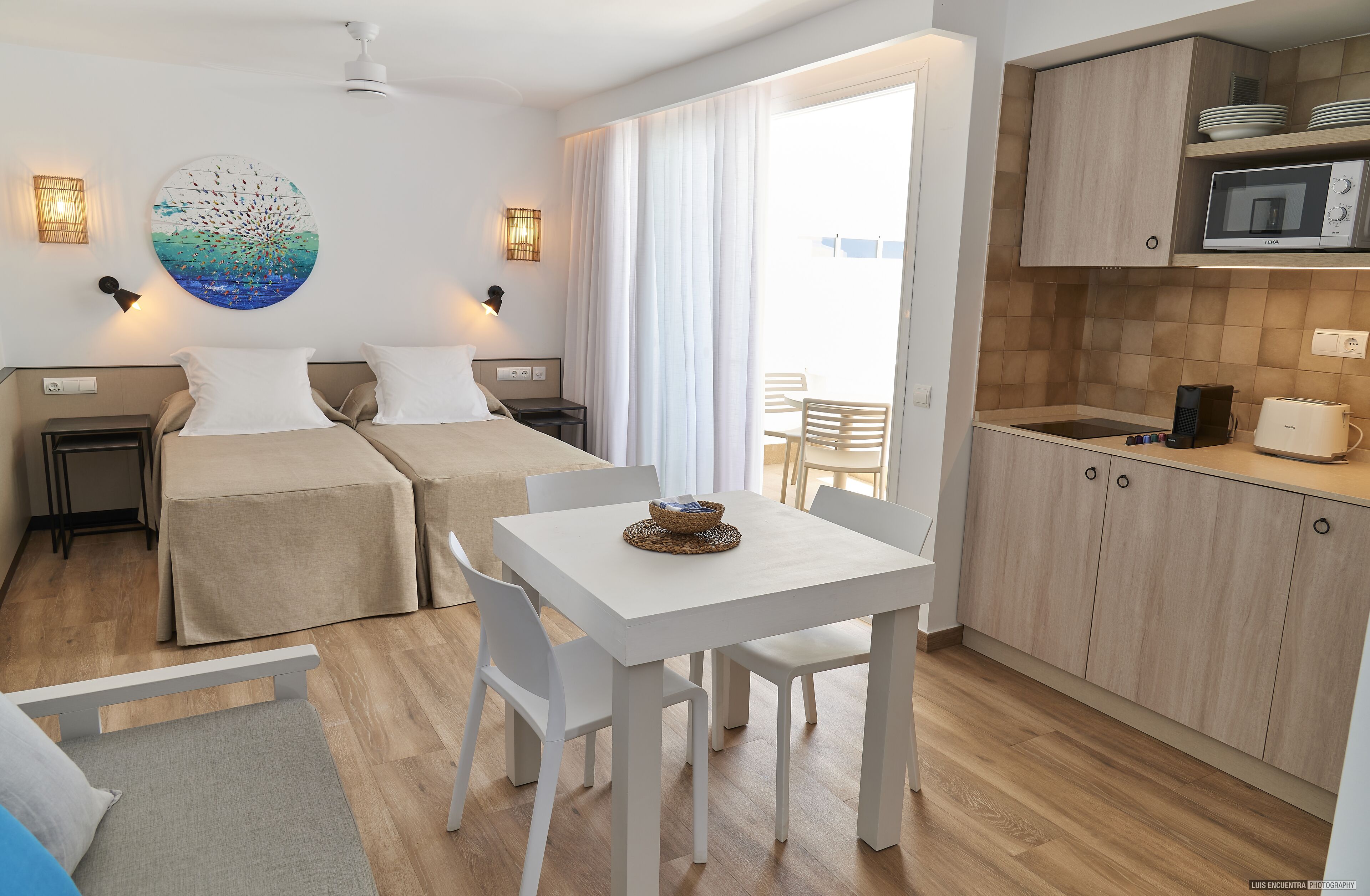 A modern hotel room with two beds, a dining table, a kitchen area, and a large window.