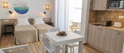 A modern hotel room with two beds, a dining table, a kitchen area, and a large window.