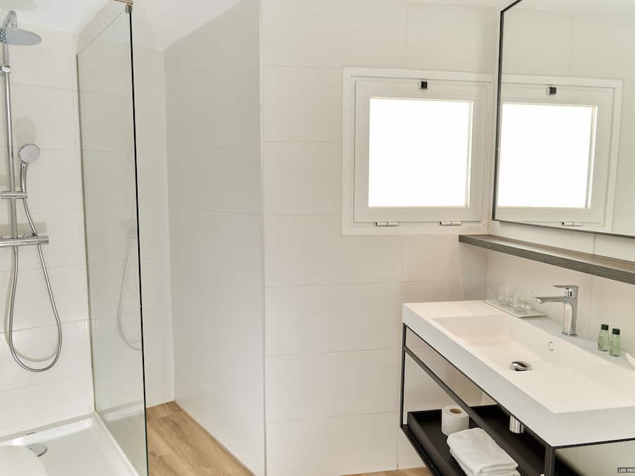 Un baño moderno con ducha de vidrio, un lavamanos blanco con espejo y una repisa con artículos de aseo.