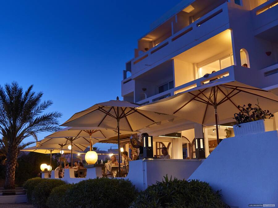 A modern hotel with outdoor seating under umbrellas, a palm tree, and a well-lit balcony area.
