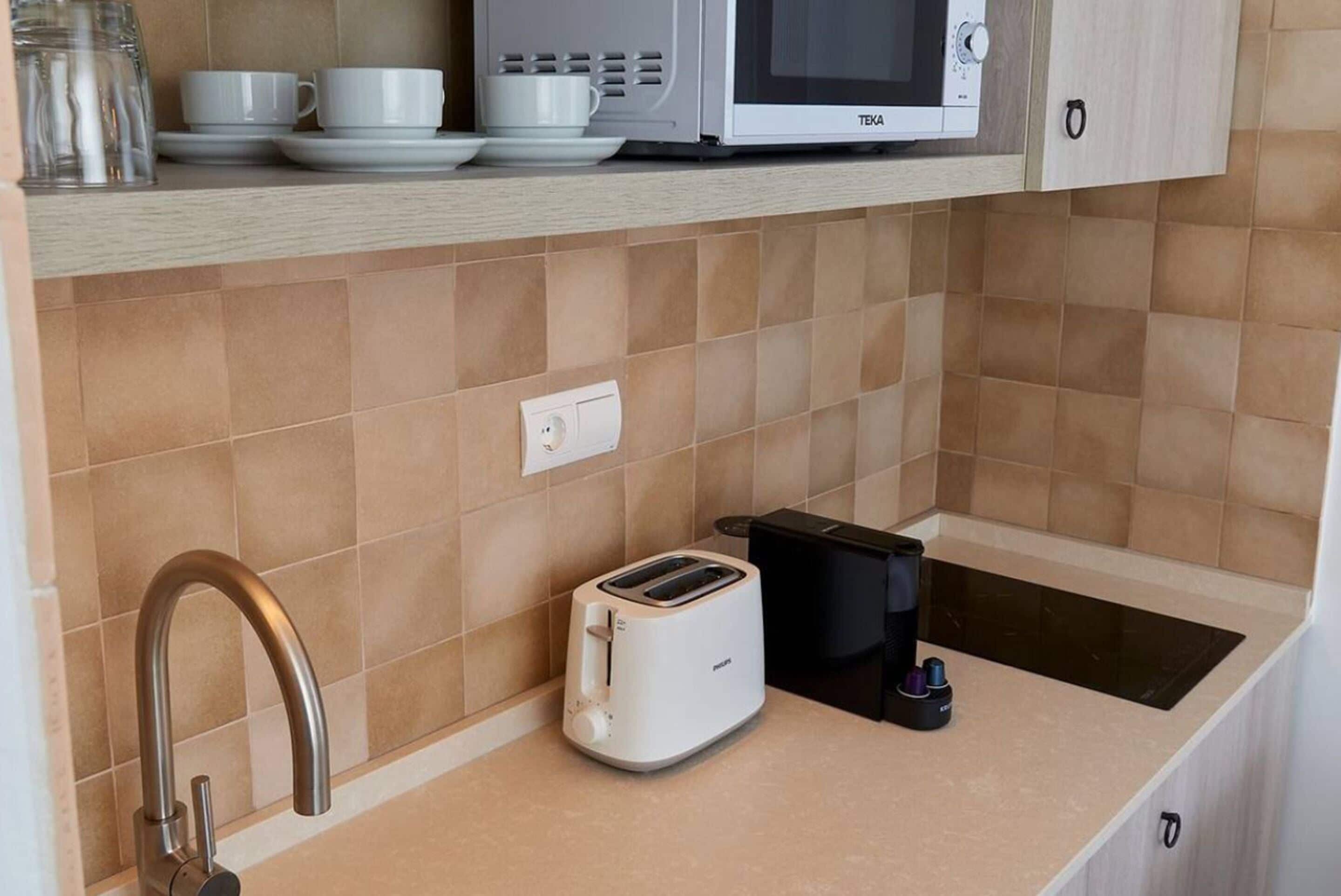 Una kitchenette con microondas, tostadora y cafetera sobre una encimera, un lavabo con grifo moderno y un armario encima de la encimera.