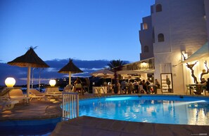 Zona de piscina con sillones, sombrillas y un bar o área de lounge con gente.