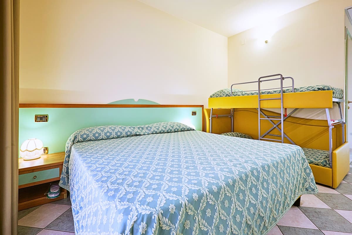 A room with a single bed, a bunk bed, a bedside table with a lamp, and a small drawer.