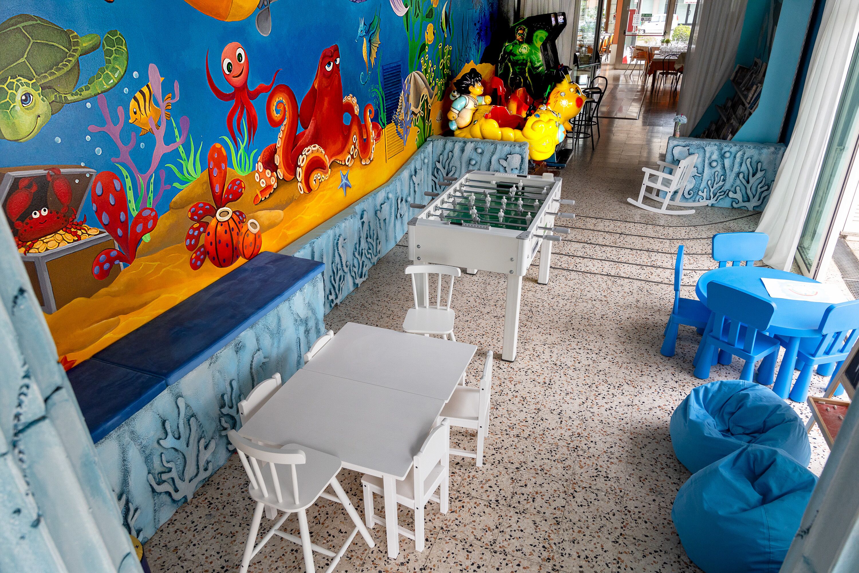 A children's play area with a mural, tables, chairs, and a foosball table.