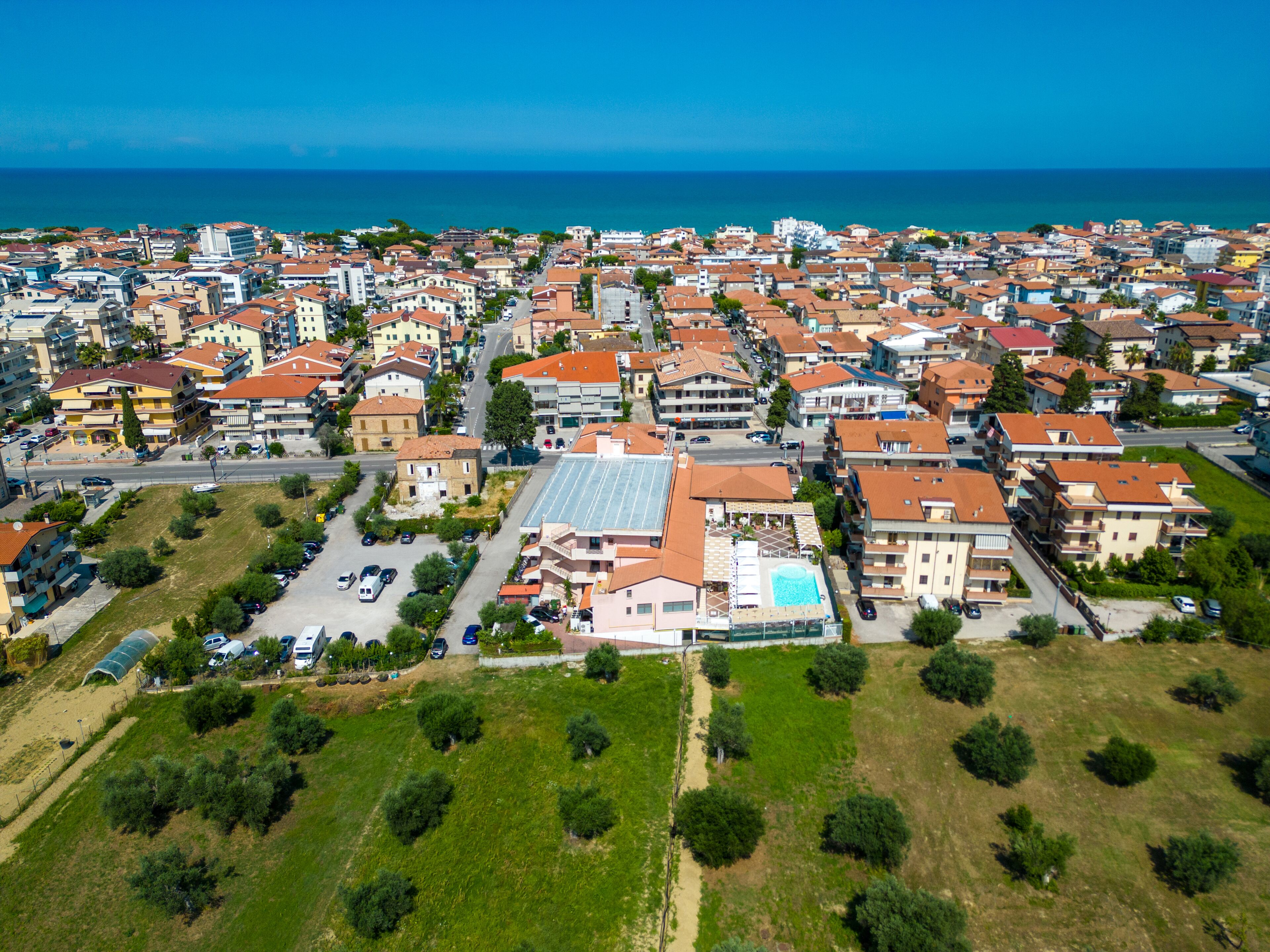 An aerial view of a coastal town with a mix of residential buildings, a prominent hotel with a swimming pool, and a clear view of the sea.