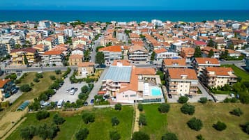 An aerial view of a coastal town with a mix of residential buildings, a prominent hotel with a swimming pool, and a clear view of the sea.