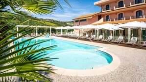 A pool area with lounge chairs and umbrellas, a building with balconies, and a clear sky. - Hotel Villa Elena (Tortoreto)