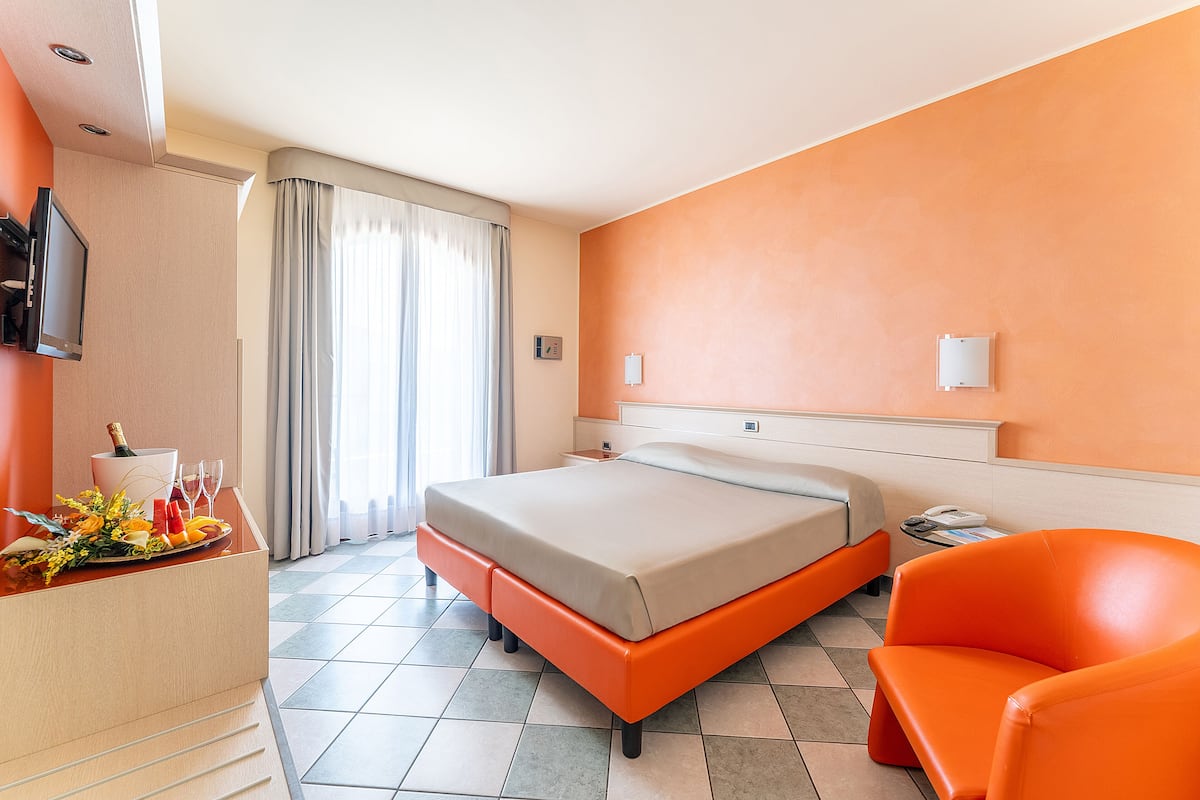 A hotel room with a bed, orange chair, desk with a bottle of champagne, glasses, and a fruit platter.