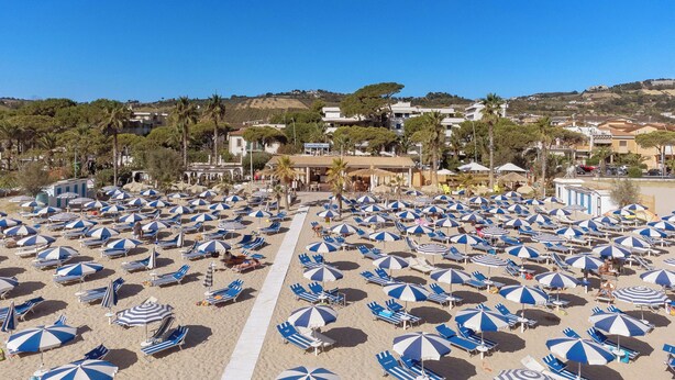 A beach with numerous blue and white striped umbrellas and sun loungers, a clear sky, and buildings in the background. - Hotel Villa Elena (Tortoreto)