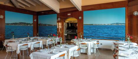 Un restaurante con mesas dispuestas para comer, amplias ventanas con vista al mar y vigas de madera en el techo.