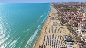 An aerial view of a coastal town with a beach lined with rows of umbrellas and sun loungers, a clear blue sea, and buildings along the shore. - Hotel Villa Elena (Tortoreto)