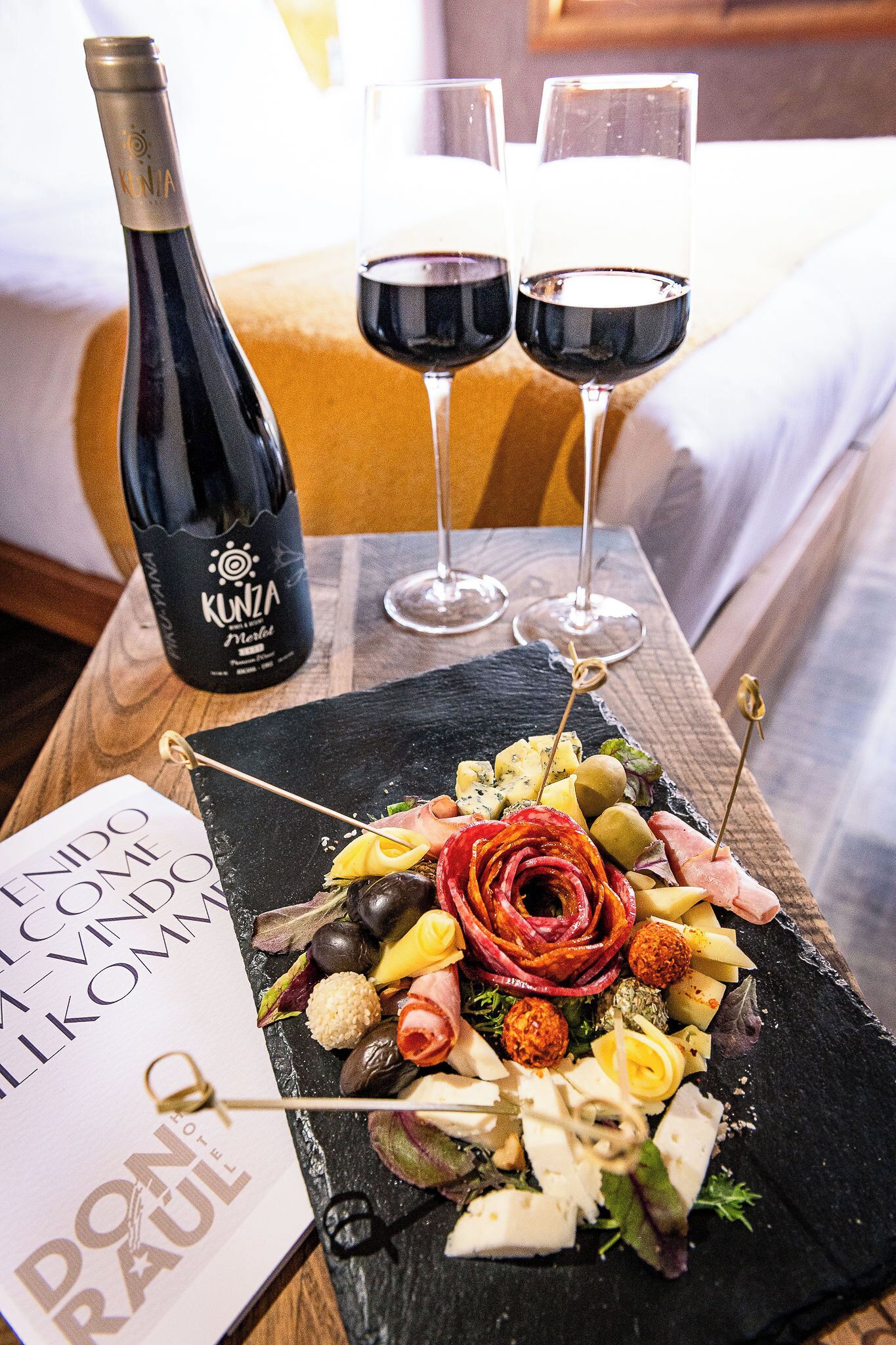 A hotel room with a wooden table displaying a charcuterie board, a bottle of wine, and two glasses of red wine.