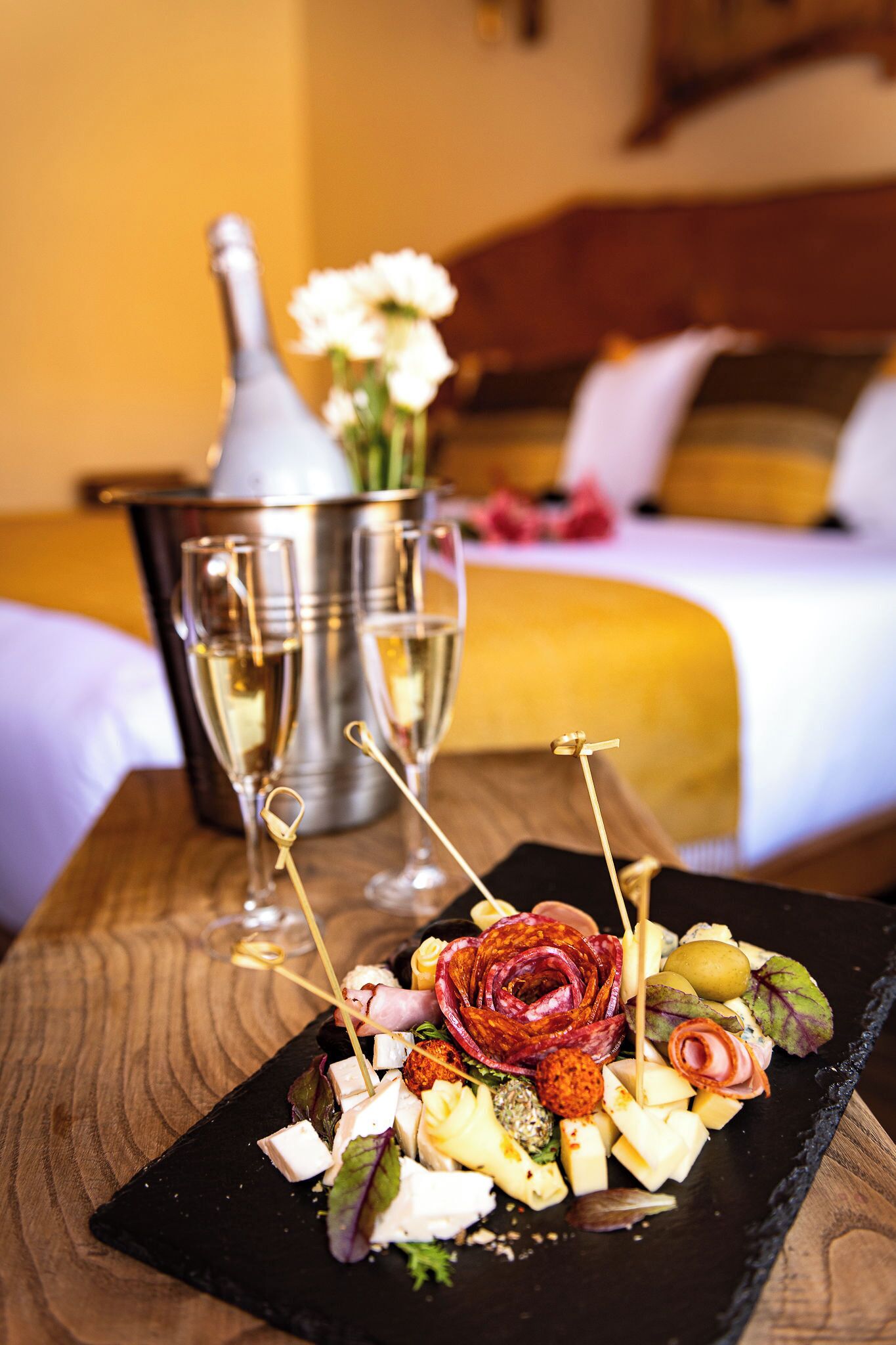 Un plateau de desserts comprenant divers fromages, des fruits et des fleurs, présenté sur une assiette d’ardoise noire, accompagné de deux coupes à champagne et d’un seau en argent avec des fleurs en arrière-plan.
