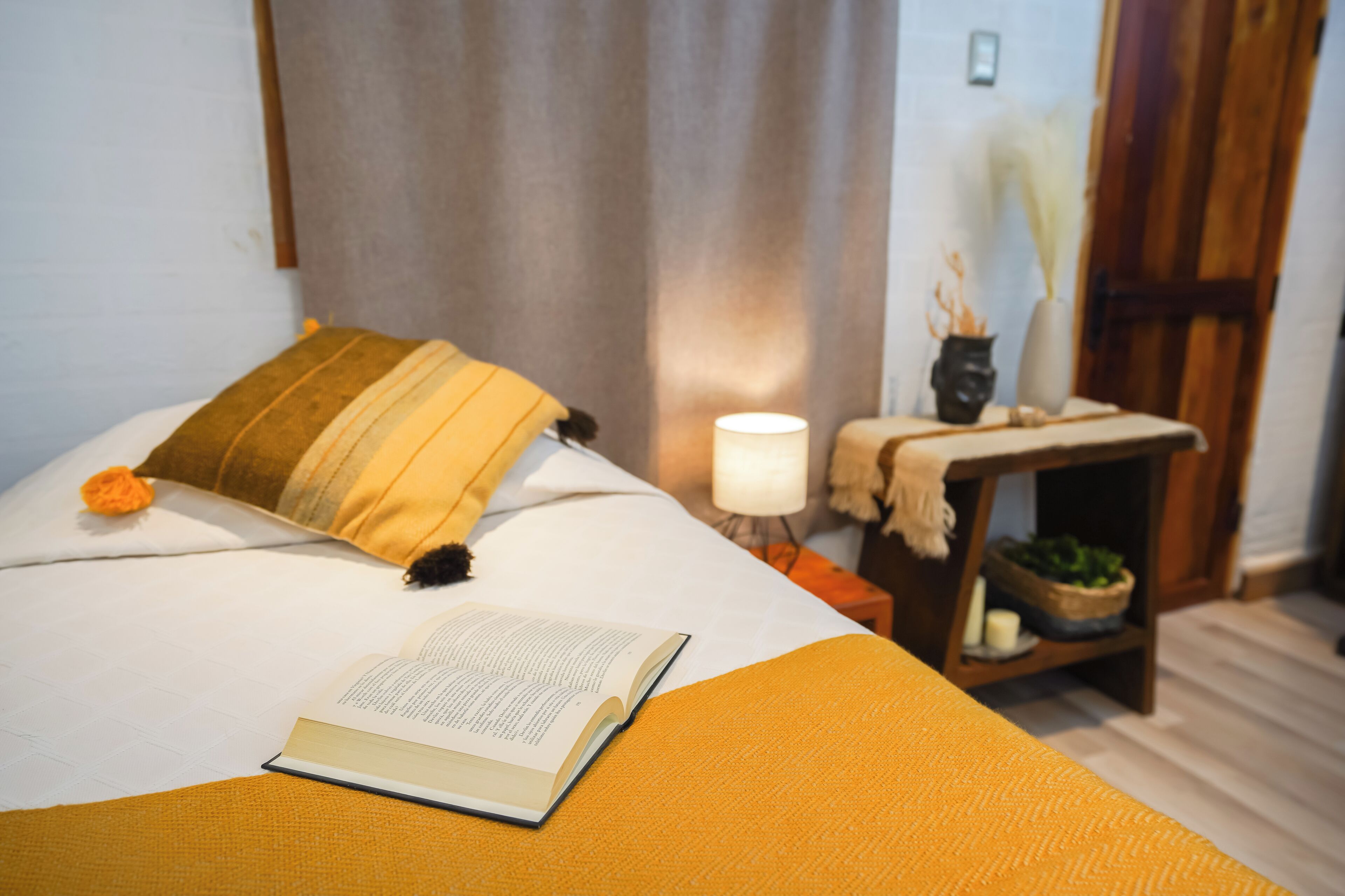 A neatly made bed with a book, a lamp, and a small table with a plant.