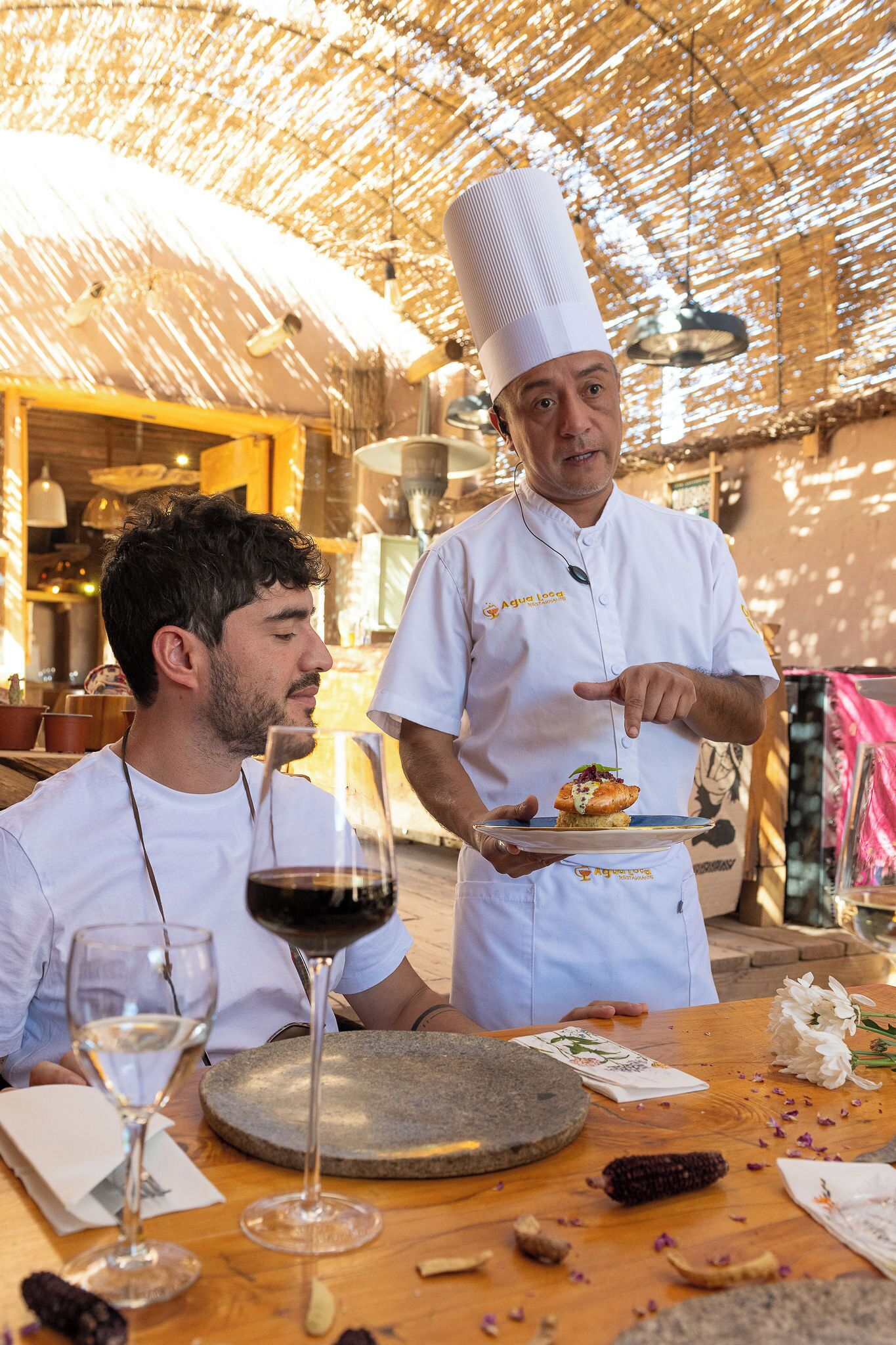 Un chef sirviendo comida a un huésped en un comedor al aire libre con una mesa de madera rústica, una copa de vino y un mortero de piedra.