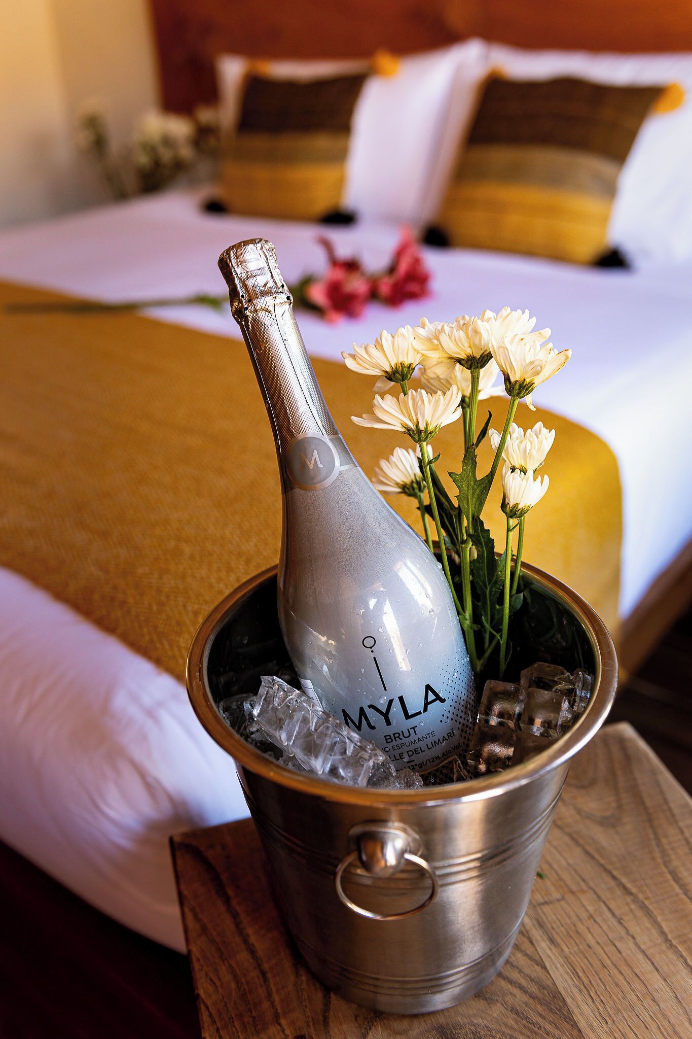 A bottle of champagne in a metal bucket with ice, placed on a wooden table in a hotel room with a bed in the background.