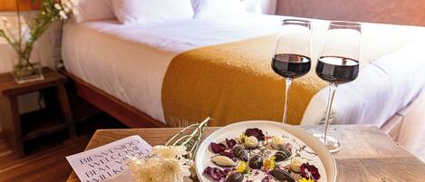 Una cama bien hecha con sábanas blancas y un edredón amarillo, una mesita de noche con un florero y una mesa con un plato de bombones variados y dos copas de vino tinto.