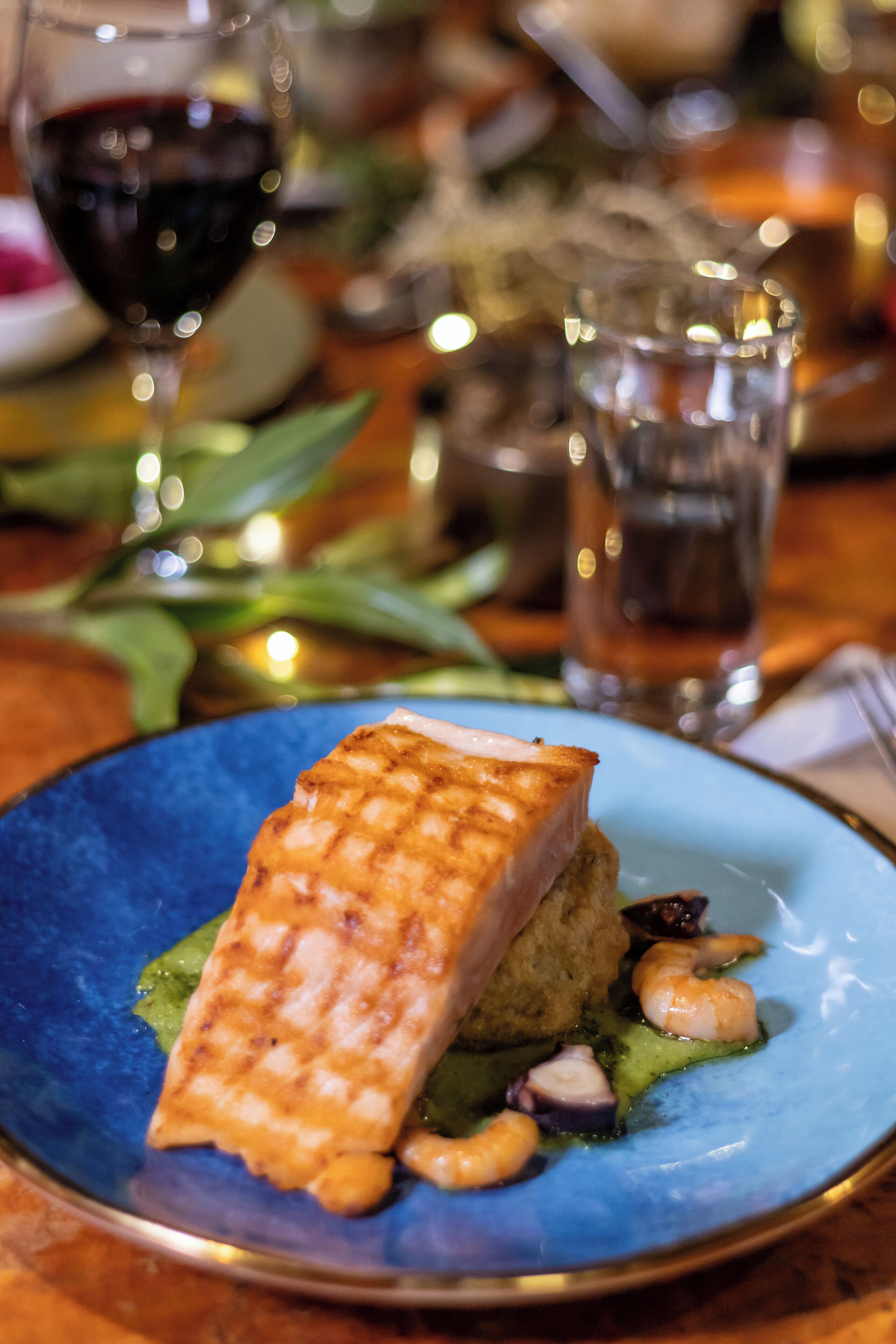 A plate with grilled fish, shrimp, and greens, accompanied by a glass of red wine and a clear glass.