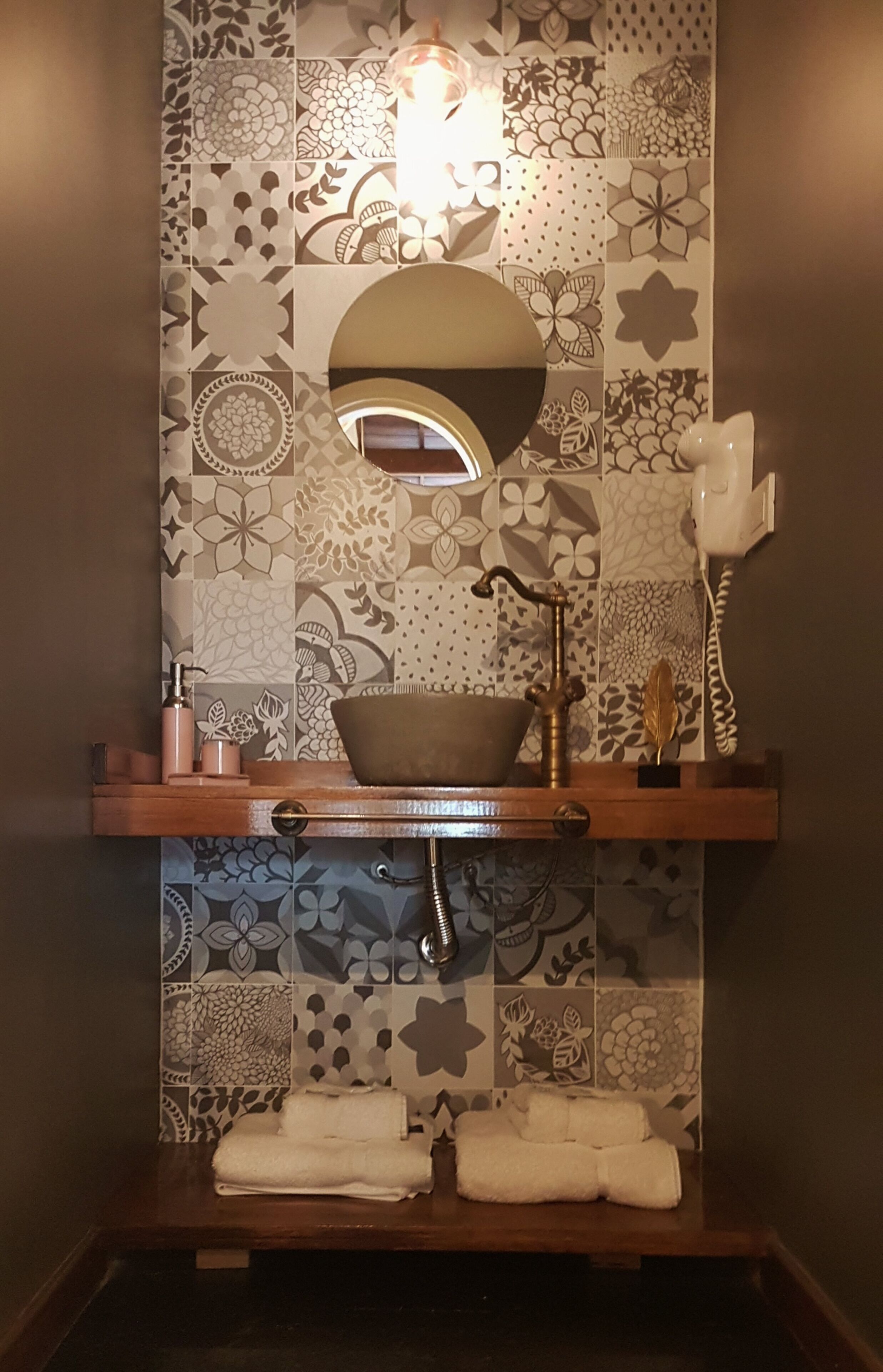 Un baño con salpicadero de azulejos estampados, un espejo redondo, un tocador de madera, un lavabo con grifo y un perchero para toallas.