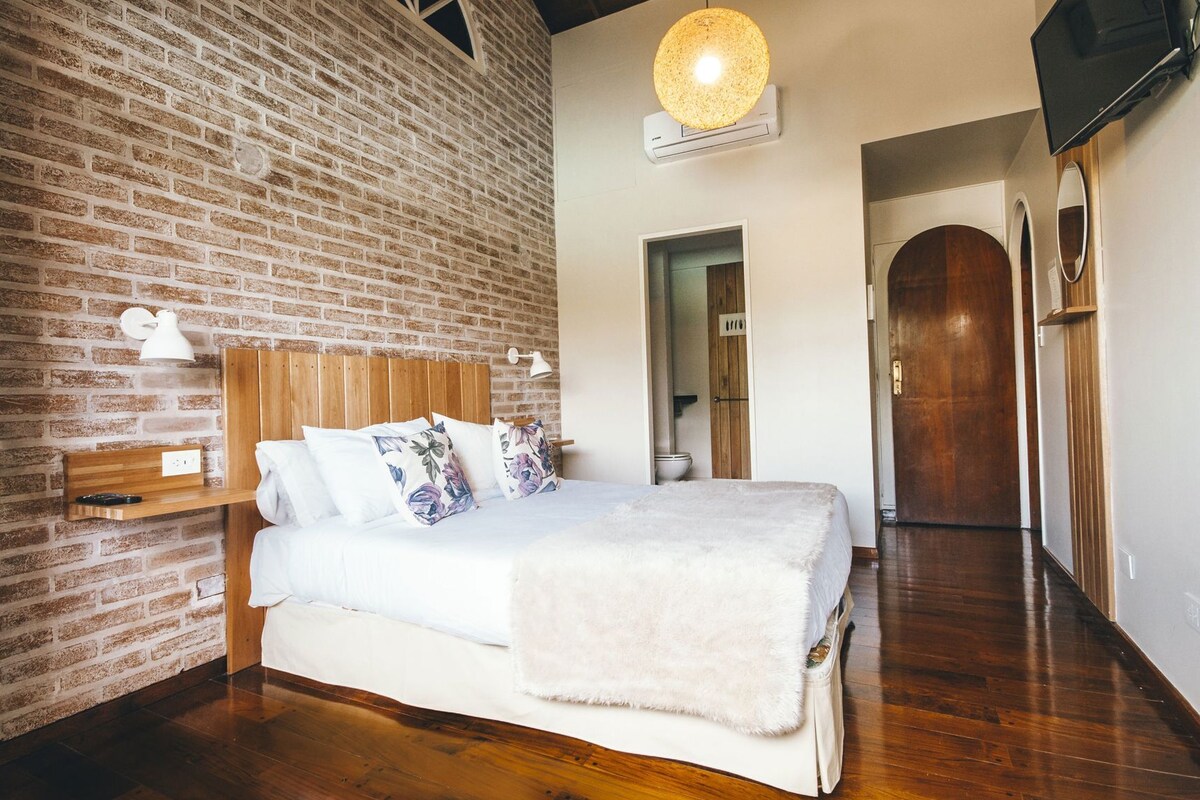 Un dormitorio con pared de ladrillo, cabecera de madera, cama con ropa blanca, lámpara colgante y suelo de madera.