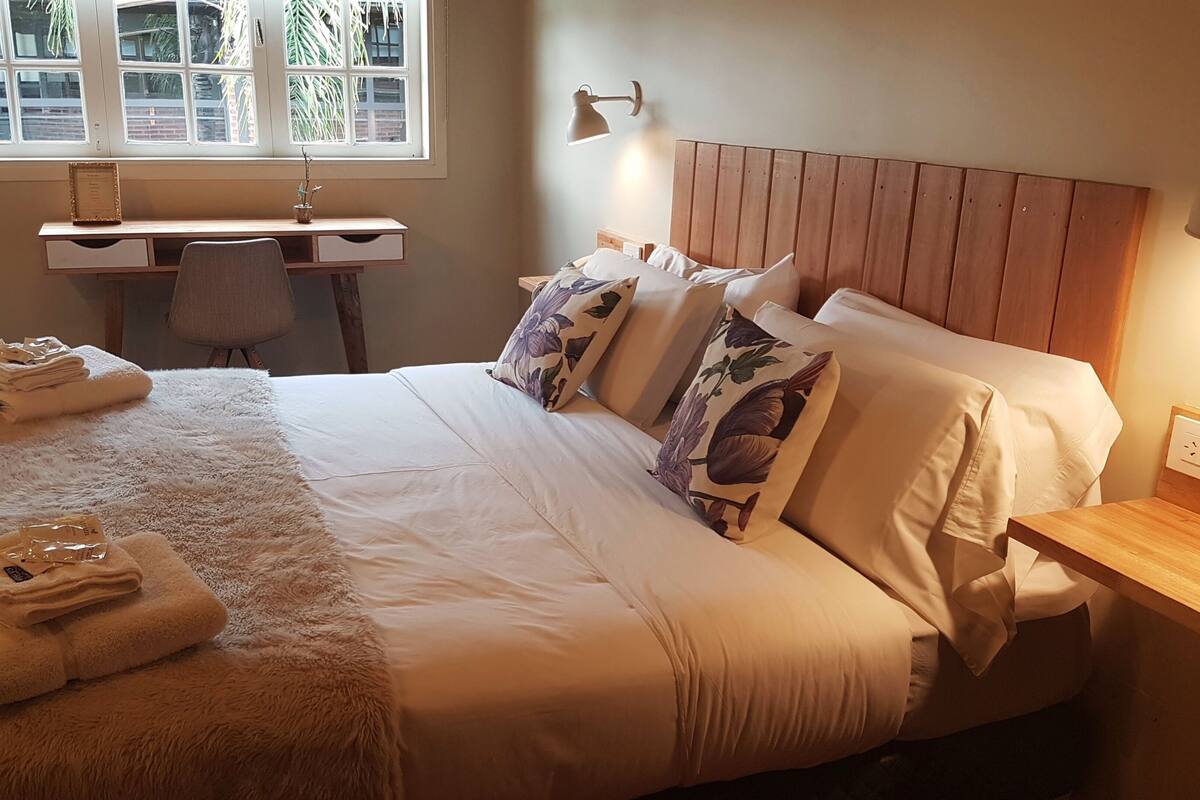 A neatly made bed with fluffy white bedding, a wooden headboard, and floral pillows. There is a desk with a chair and a lamp, and a window with blinds.