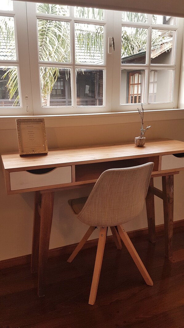 Un escritorio de madera con una silla de fibras tejidas, un certificado enmarco y un pequeño jarrón con una rama.