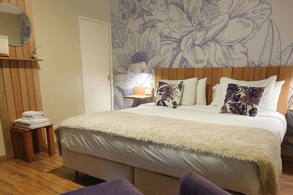 Un dormitorio con una cama grande, cabecera de madera, un mural floral, una mesita de noche pequeña y dos sillas moradas.
