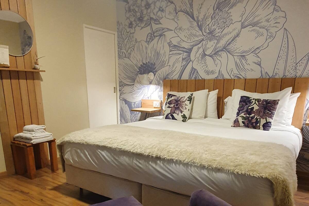 A bedroom with a large bed, wooden headboard, and a floral wall mural. There are two purple armchairs and a small wooden table with towels.