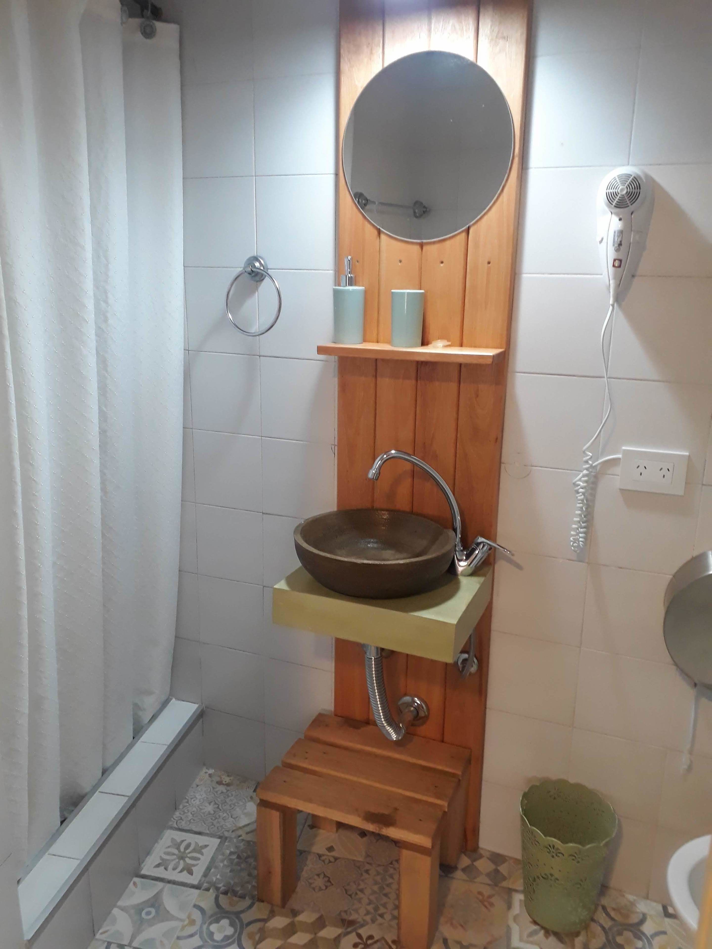 A bathroom with a wooden vanity, a round mirror, a vessel sink, a handheld showerhead, a hairdryer, and a green trash bin.