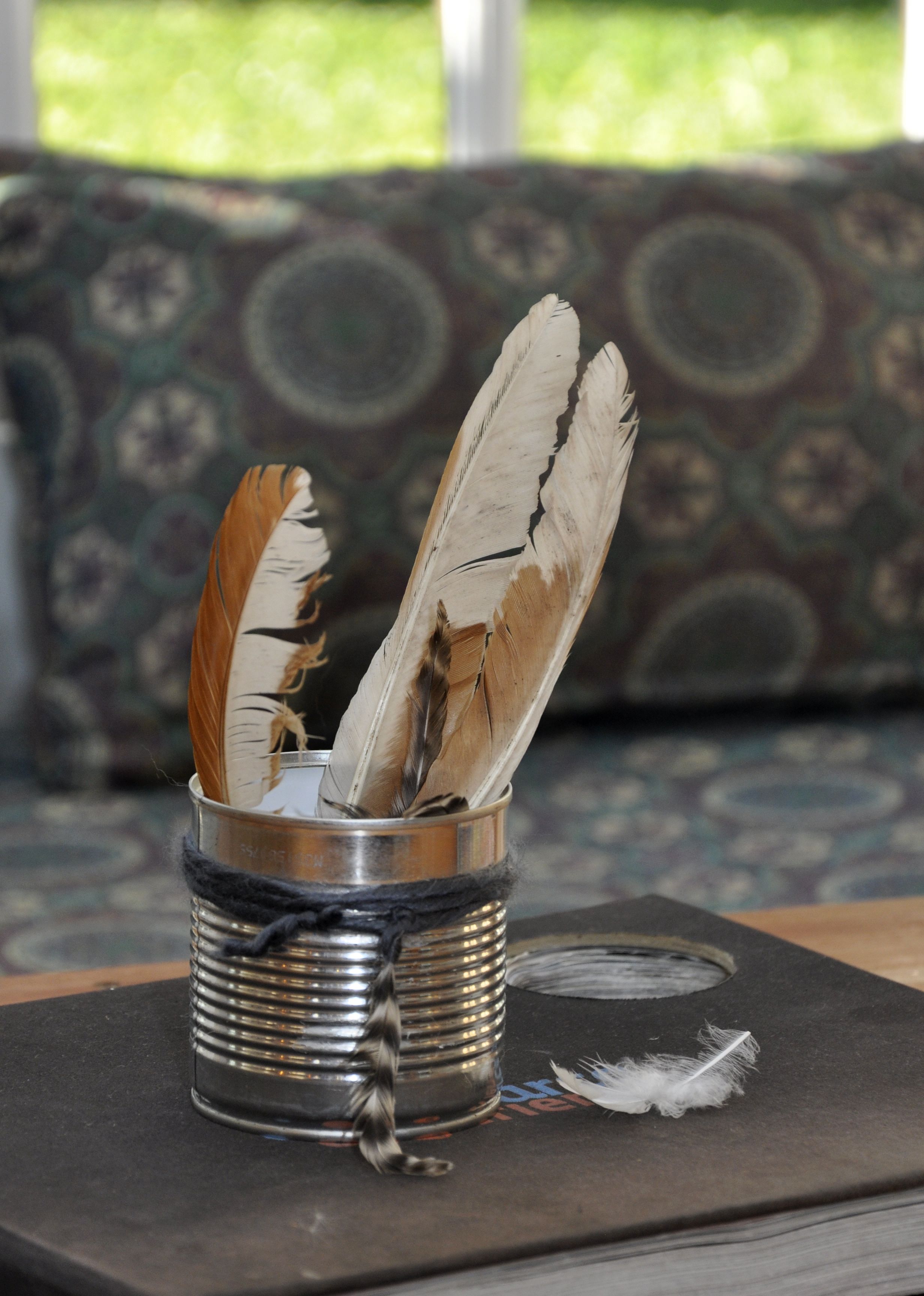 Un recipiente metálico con plumas sobre una superficie oscura, posiblemente un libro o cuaderno, con un cojín estampado al fondo.