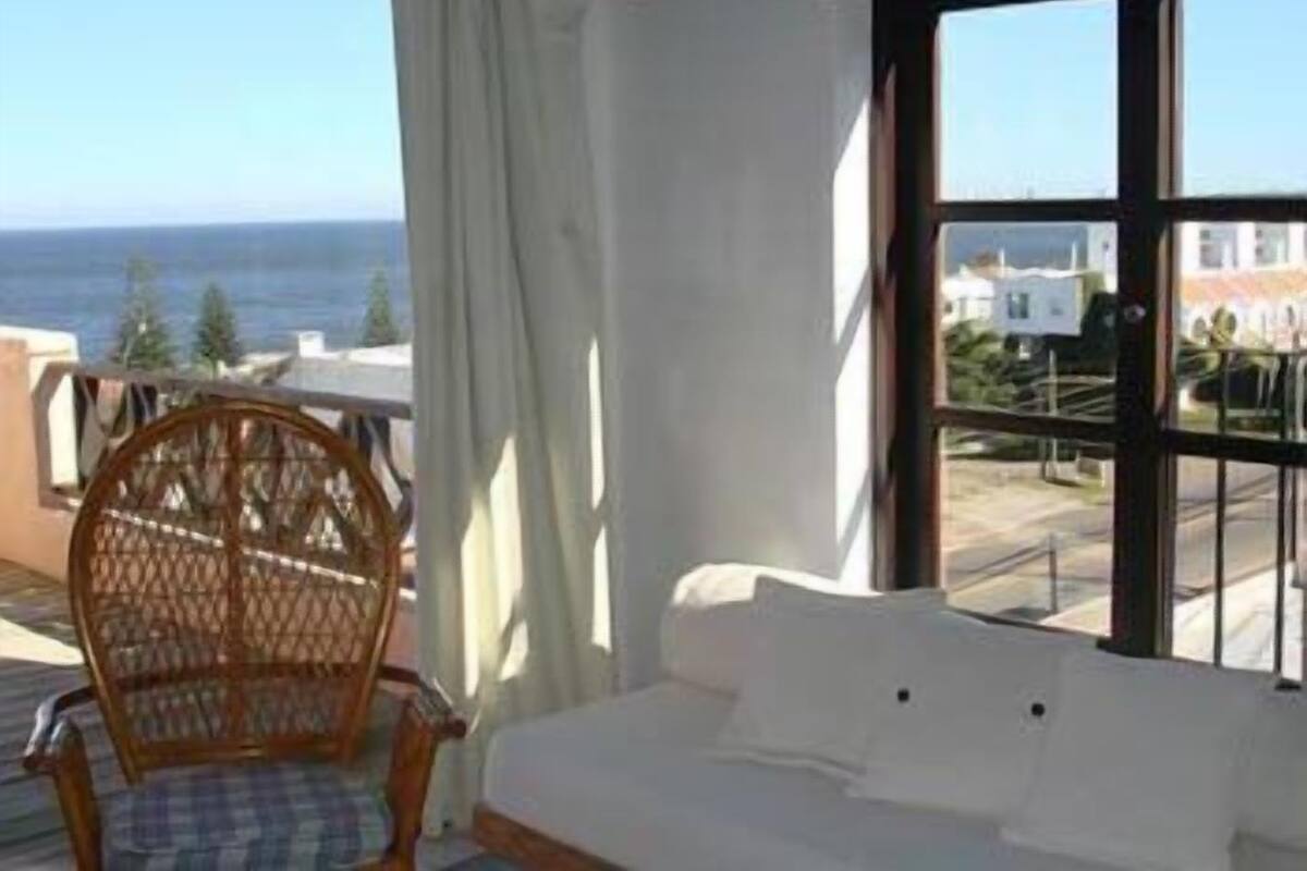 A balcony with a wicker chair and a white sofa, overlooking a coastal view.