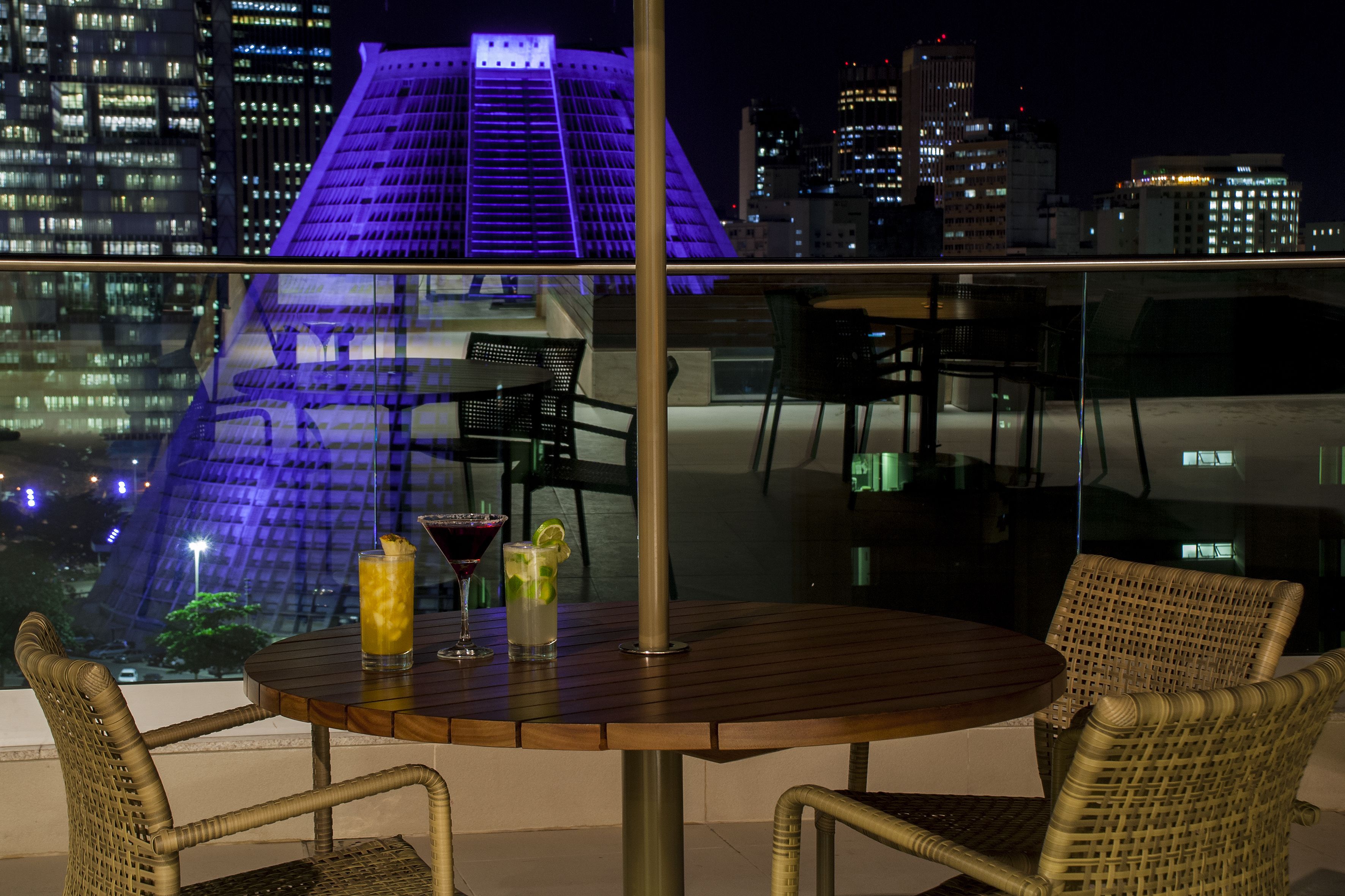 A rooftop bar with a view of a city skyline at night, featuring a modern building with illuminated blue lights.