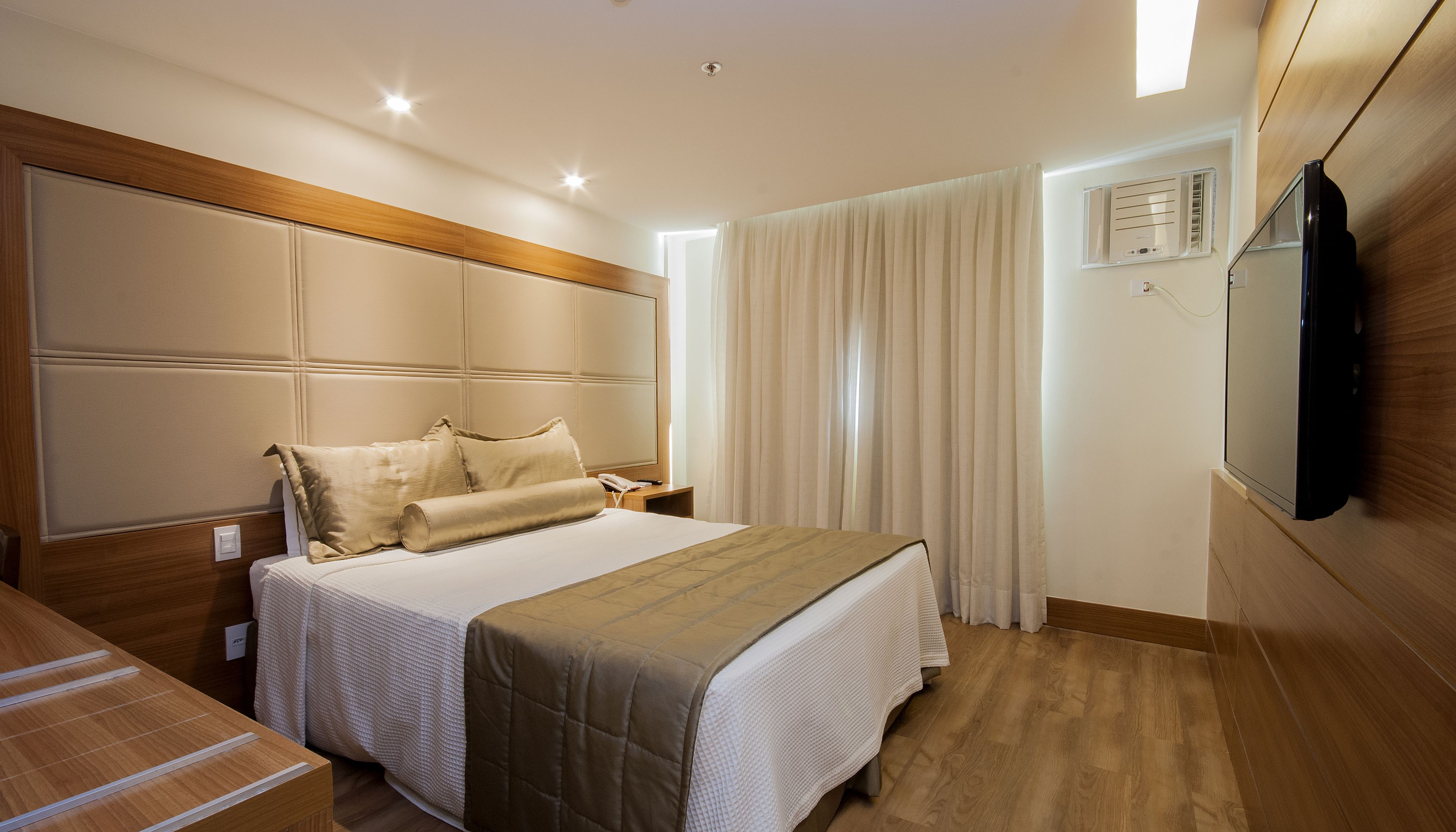 Habitación de hotel con una cama grande, cabecera de madera, mesita de noche con lámpara y televisor montado en la pared.
