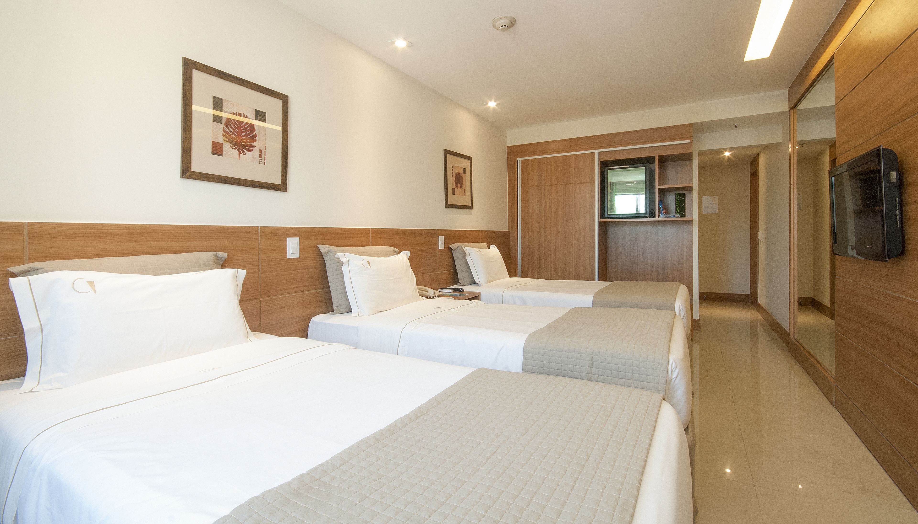 Quarto de hotel com três camas, cabeceira de madeira, televisão de tela plana e uma pequena cozinha compacta.