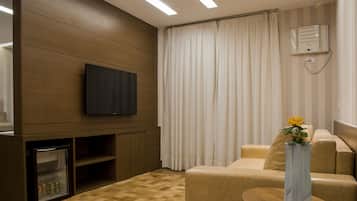 Uma sala de estar moderna com televisão de tela plana fixada em uma parede de madeira, mini-geladeira embutida, sofá bege, um vaso branco com flores e uma grande janela com cortinas brancas.
