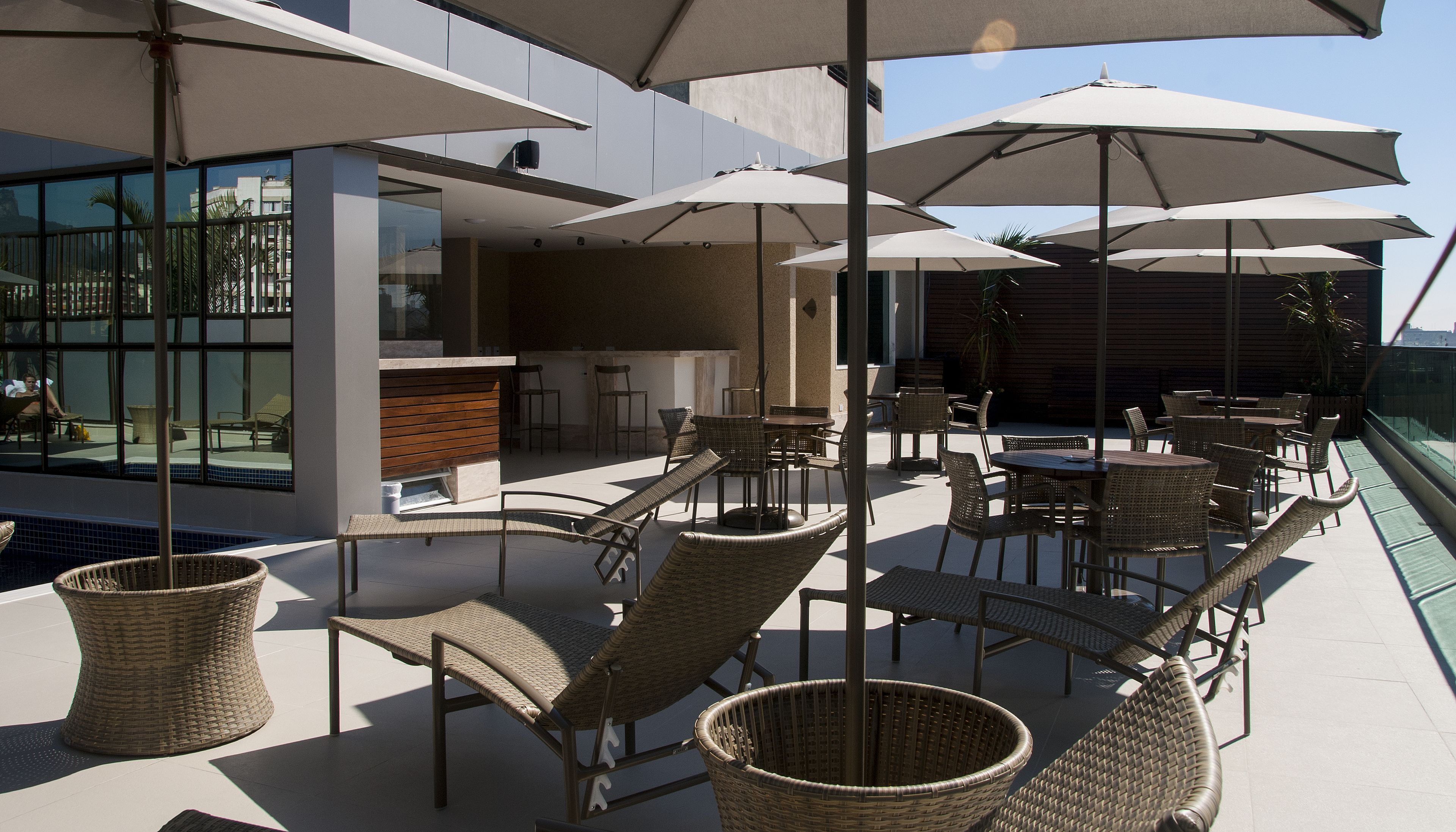 Terraza en la azotea con sillas y mesas de mimbre, y sombrillas, con vistas a una piscina y un edificio con grandes ventanales.