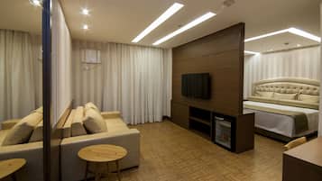 Quarto de hotel moderno com um sofá, uma pequena mesa de madeira, uma TV fixada em um painel de madeira, uma cama com cabeceira estofada e um minibar.