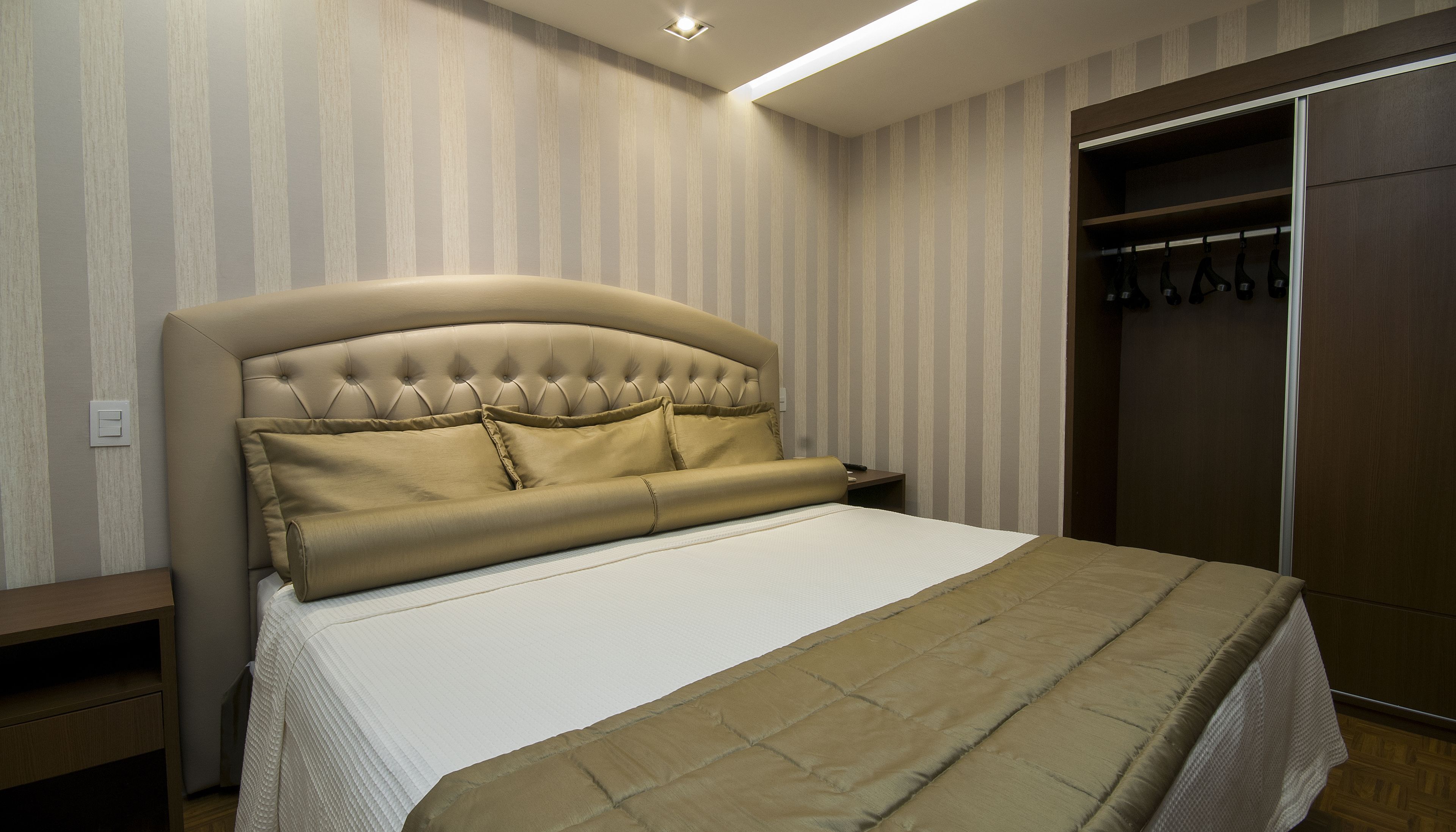 A bedroom with a tufted headboard, a bed with a beige blanket, a nightstand, and a wardrobe with hanging items.