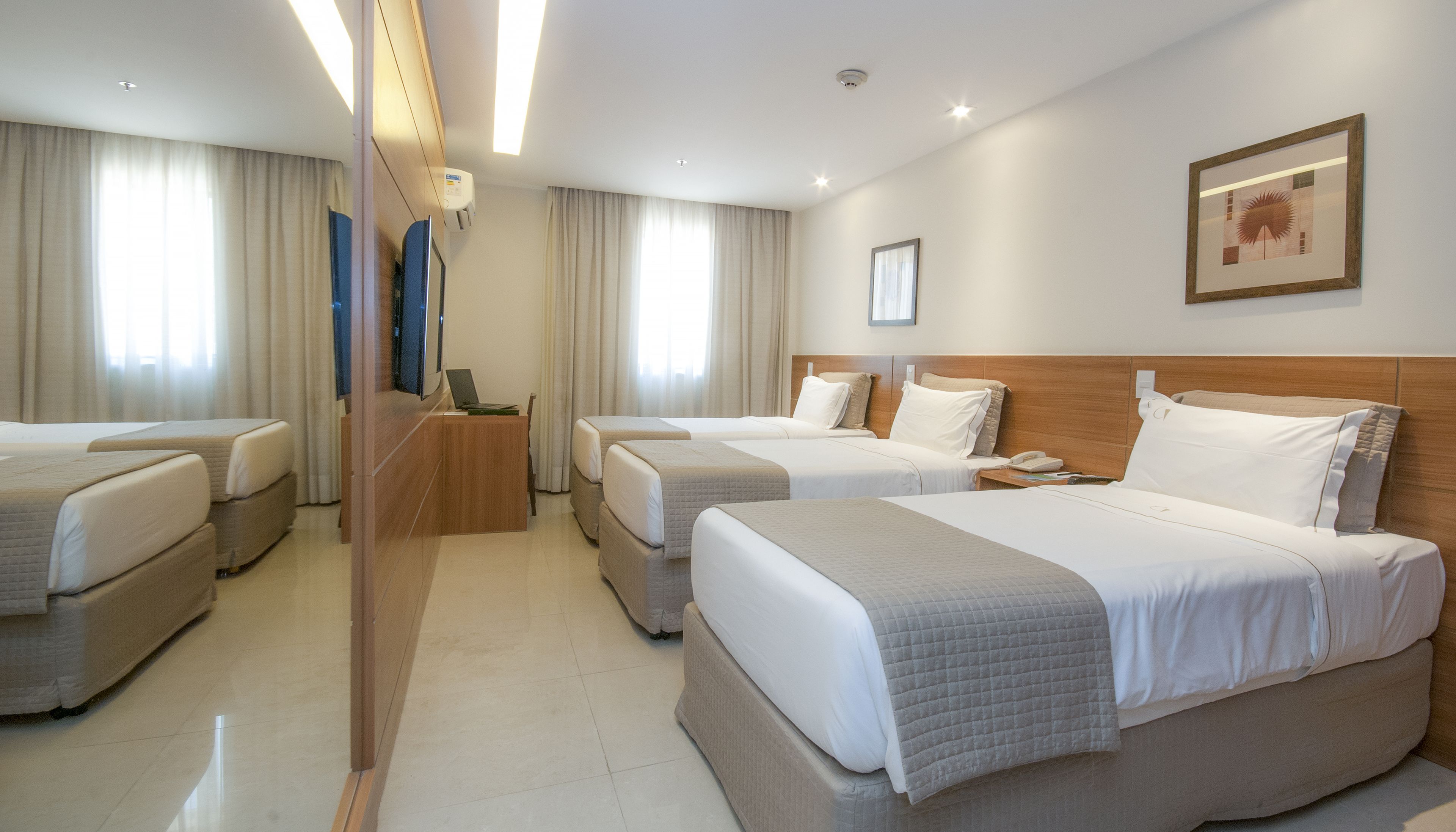 Quarto de hotel com três camas, uma escrivaninha de madeira, televisão de tela plana e um quadro na parede.